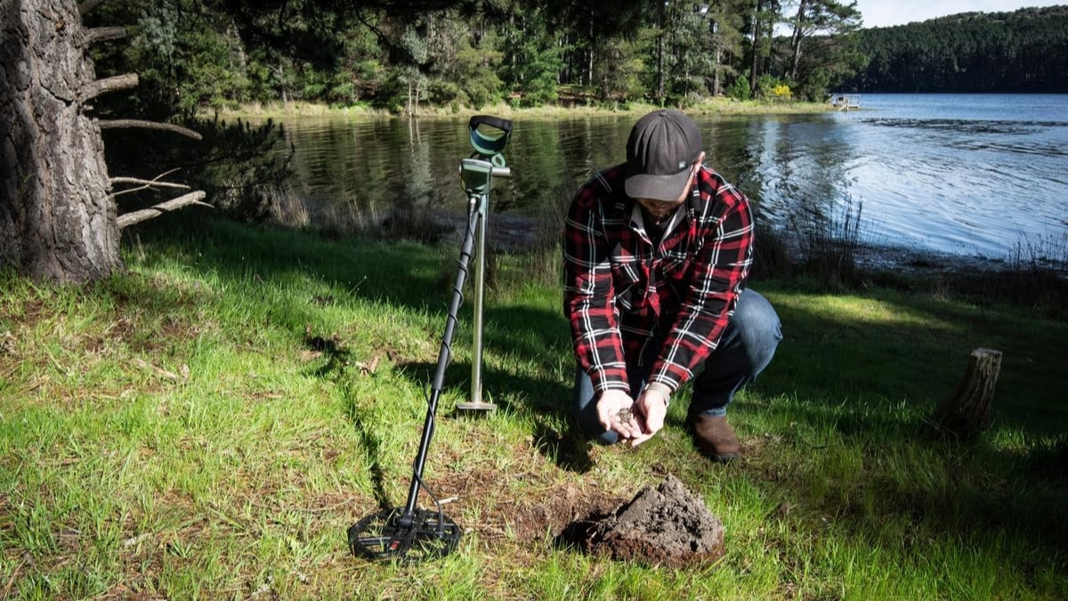 A person using a metal detector by a lake holds soil while searching for buried items.