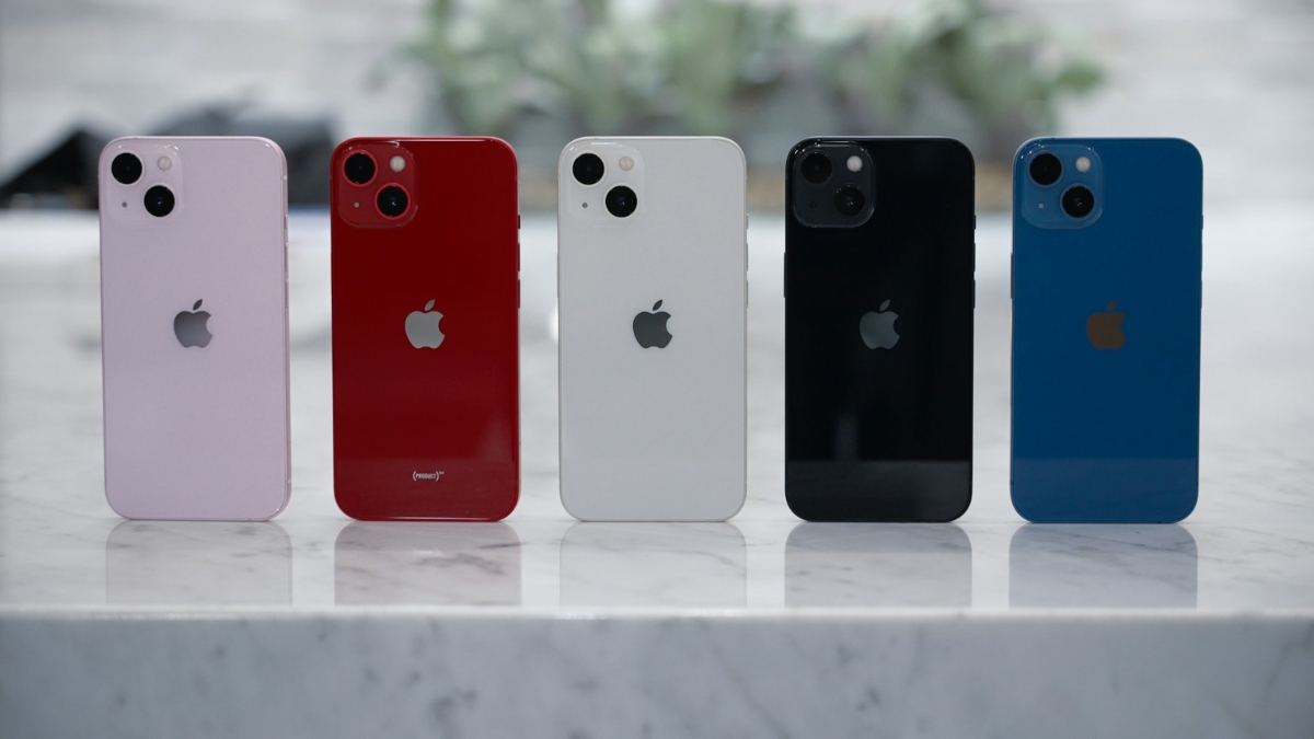 Five Apple iPhone 13 models in pink, red, Starlight, black, and blue stand upright on a marble surface.