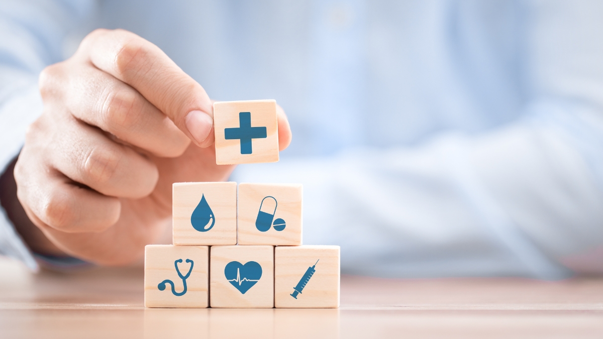 Hand stacking wooden blocks with healthcare icons, including a medical cross, heart monitor, stethoscope, syringe, drop, and pills.