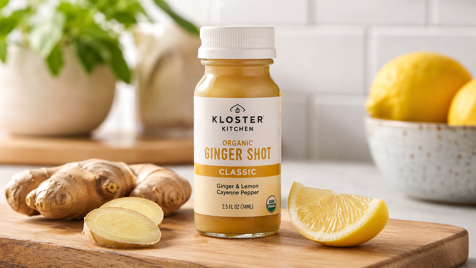 Kloster Kitchen Organic Ginger Shot Classic bottle with fresh ginger and lemon on a wooden cutting board in a bright kitchen.