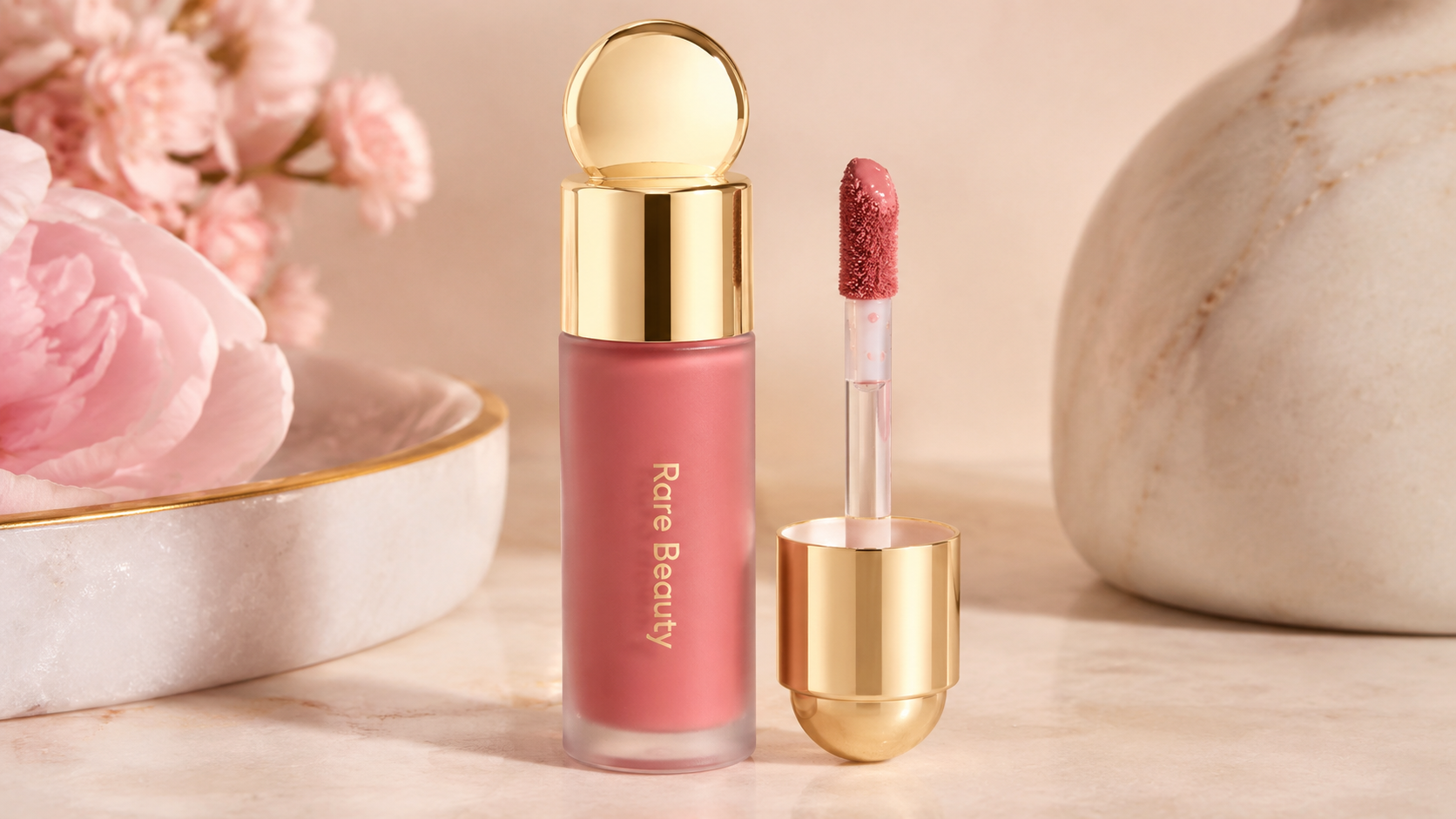 Rare Beauty Soft Pinch Liquid Blush in a frosted glass bottle with a gold cap and doe-foot applicator, styled on a marble surface with soft pink flowers in an elegant beauty editorial setting.