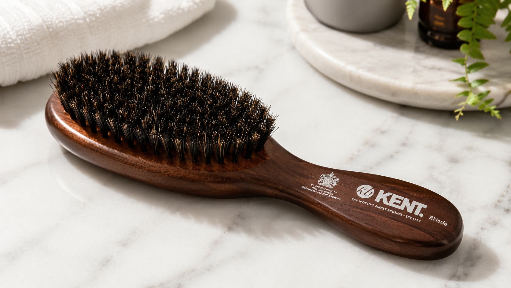 Kent Brushes Men's Oval Bristle Brush with dark wooden handle resting on a marble bathroom countertop, premium natural bristle hairbrush for men's grooming and beard care.