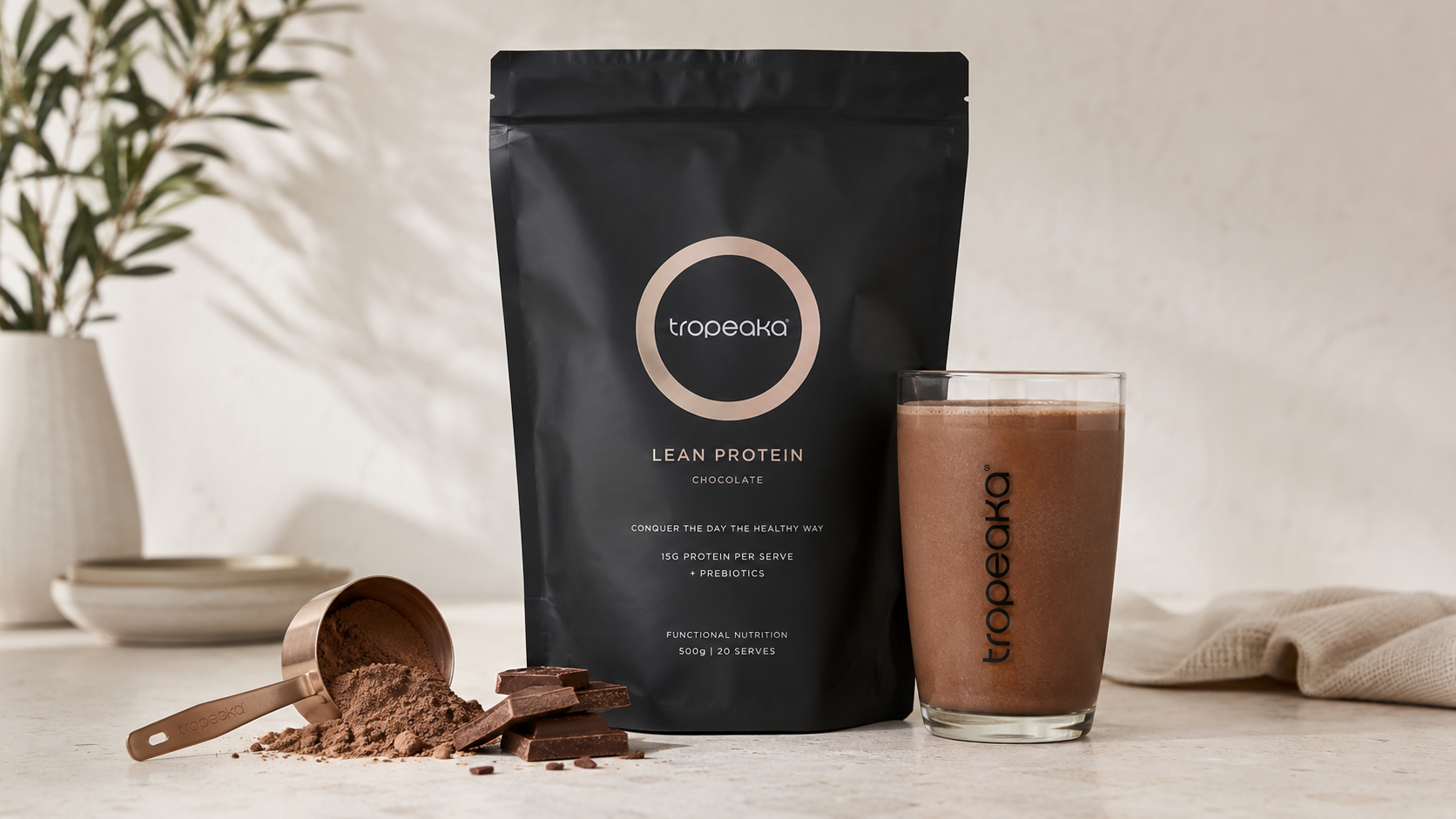 Tropeaka Lean Protein Chocolate powder in a black pouch beside a chocolate protein shake, cocoa powder, scoop, and chocolate pieces on a clean countertop with a soft neutral background.