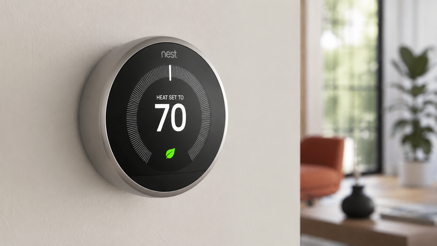 Google Nest Learning Smart Thermostat mounted on a wall displaying 70°F in a modern living room, showcasing its smart home temperature control and energy-saving design.